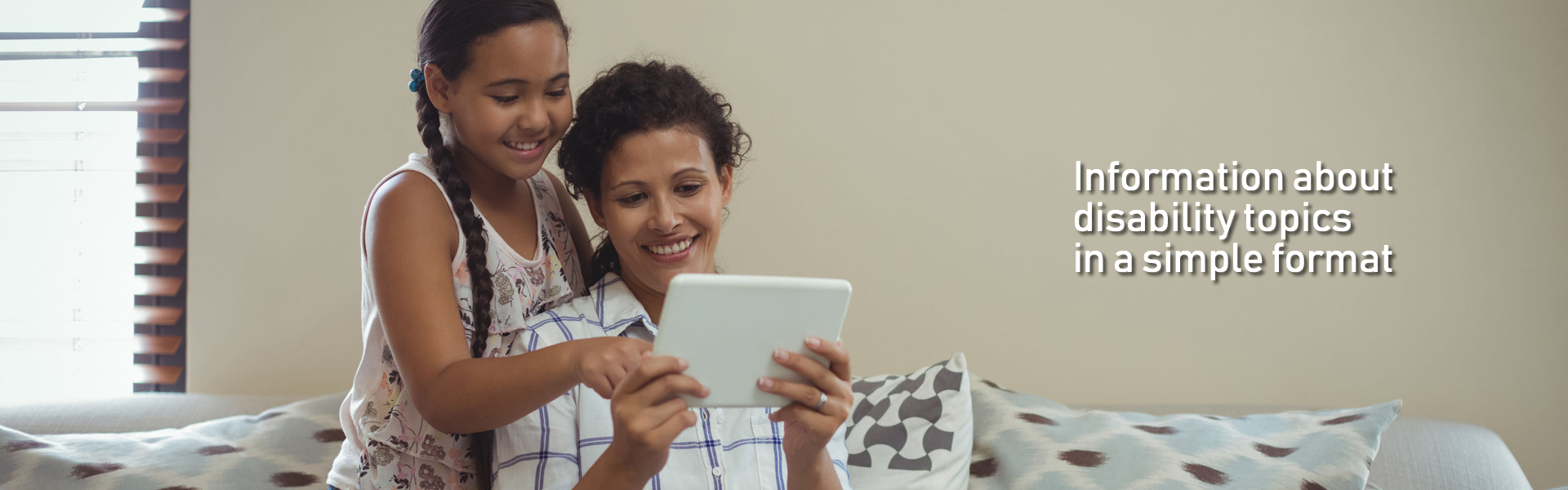 Indoors. On a sofa, a  mother and her daughter smile. The mother holds a tablet at chest height. The daughter is kneeling behind the mother's right shoulder and touches the screen of the tablet. Text at right over the image: Information about disability topics in a simple format.